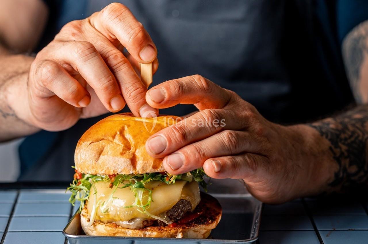 Un cuisinier assemble un burger garni de fromage fondu, de pousses vertes et de sauce sur une grille métallique.
