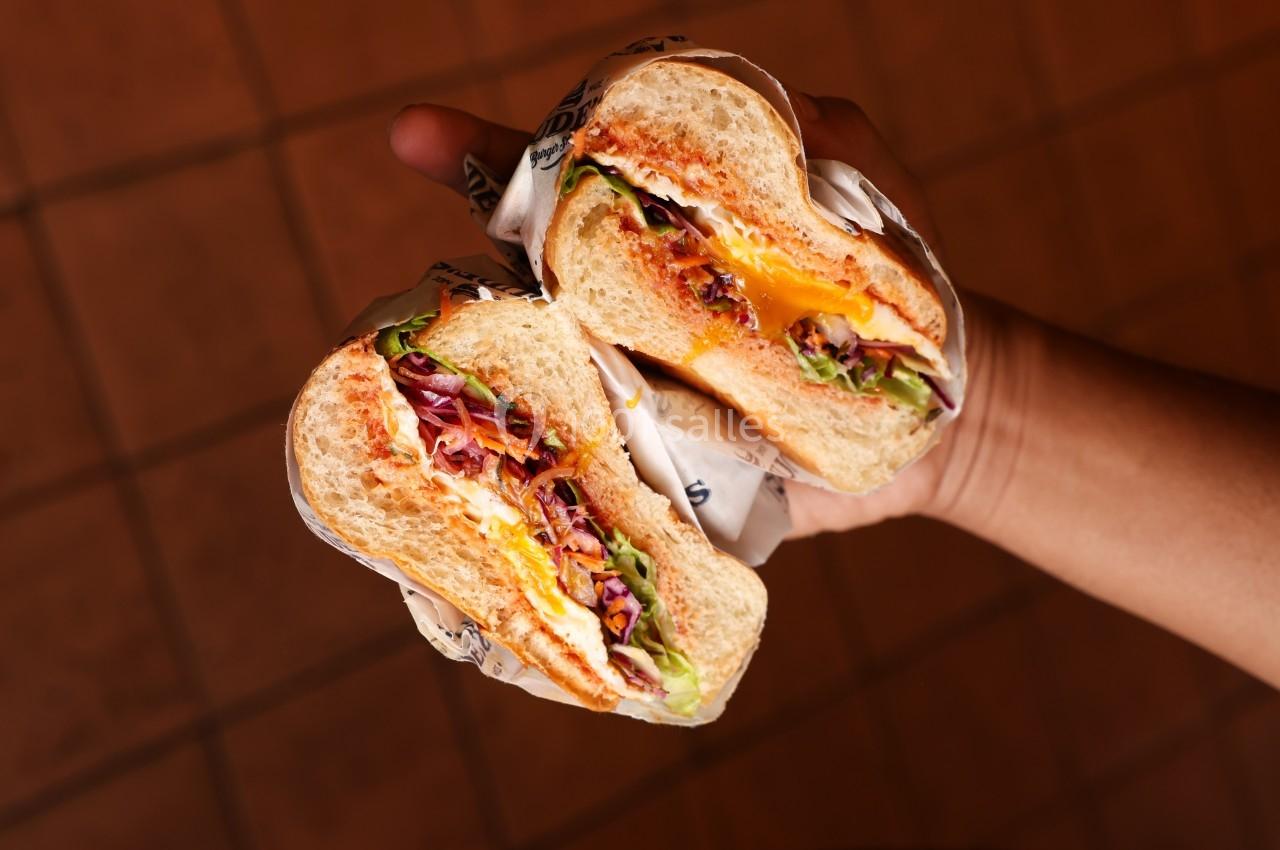 Sandwich coupé en deux, garni de fromage fondu, légumes frais et sauce, tenu dans une main sur un fond carrelé.