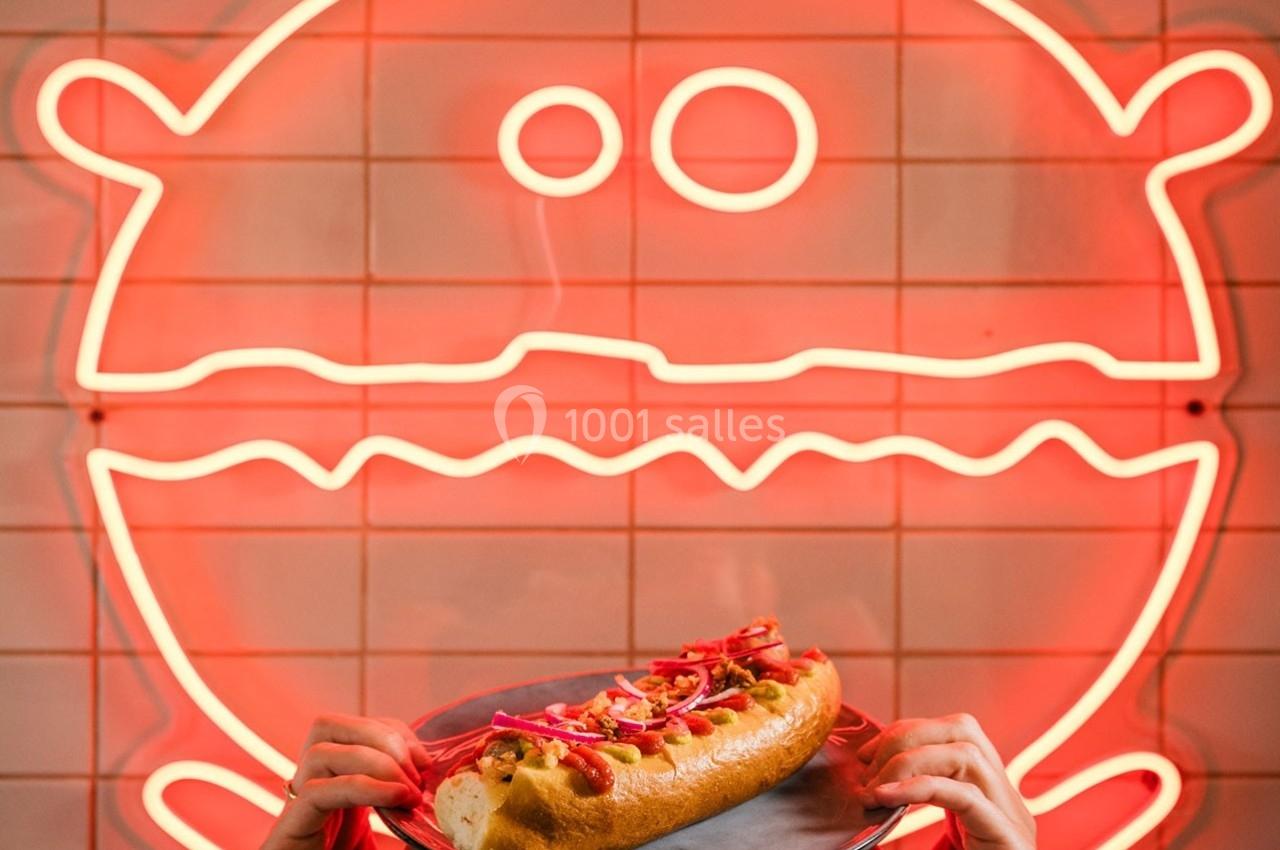 Un hot-dog garni est tenu devant un néon rouge en forme de visage stylisé de burger sur un mur carrelé.