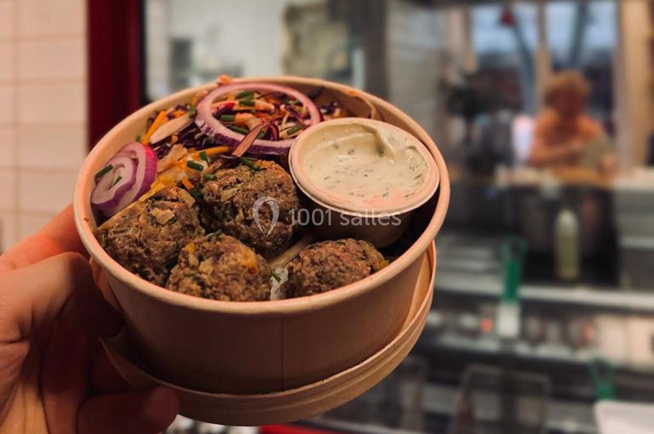 Bol en carton contenant des boulettes végétariennes, crudités colorées et sauce, tenu devant une cuisine ouverte.
