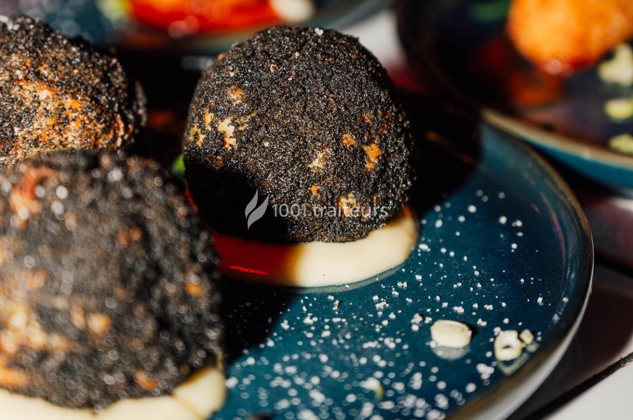Boulettes panées noires servies sur une purée, présentées sur une assiette bleue avec des garnitures.