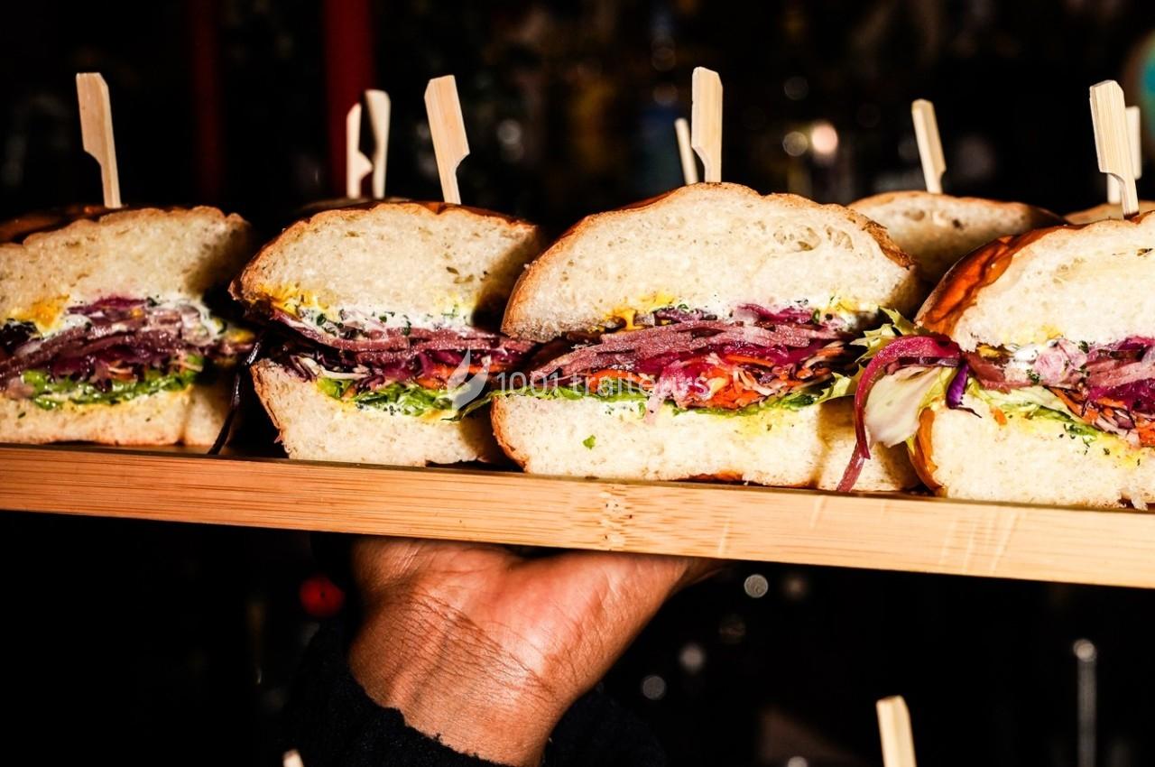 Quatre sandwichs garnis de légumes et de viande, présentés sur un plateau en bois tenu par une main.