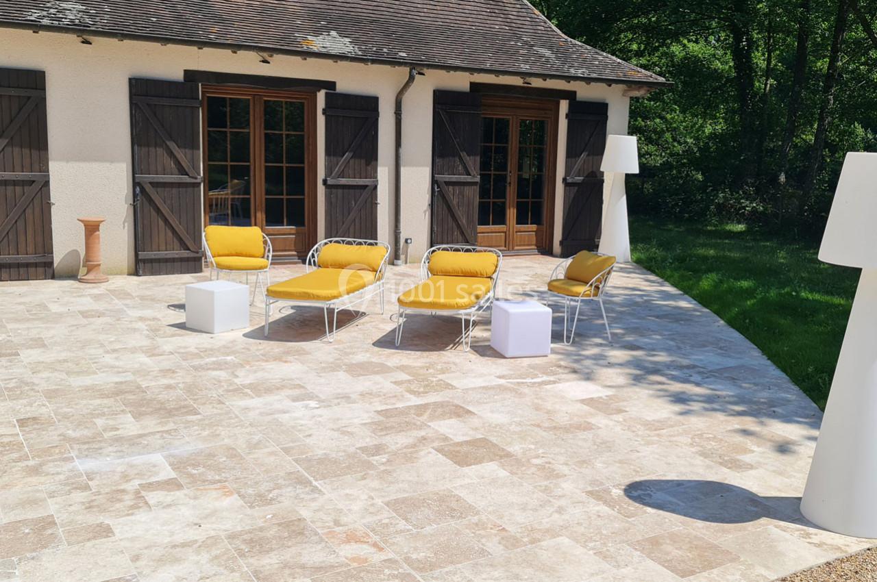 Terrasse en pierre avec mobilier de jardin jaune et blanc, devant une maison aux volets en bois et un jardin verdoyant.