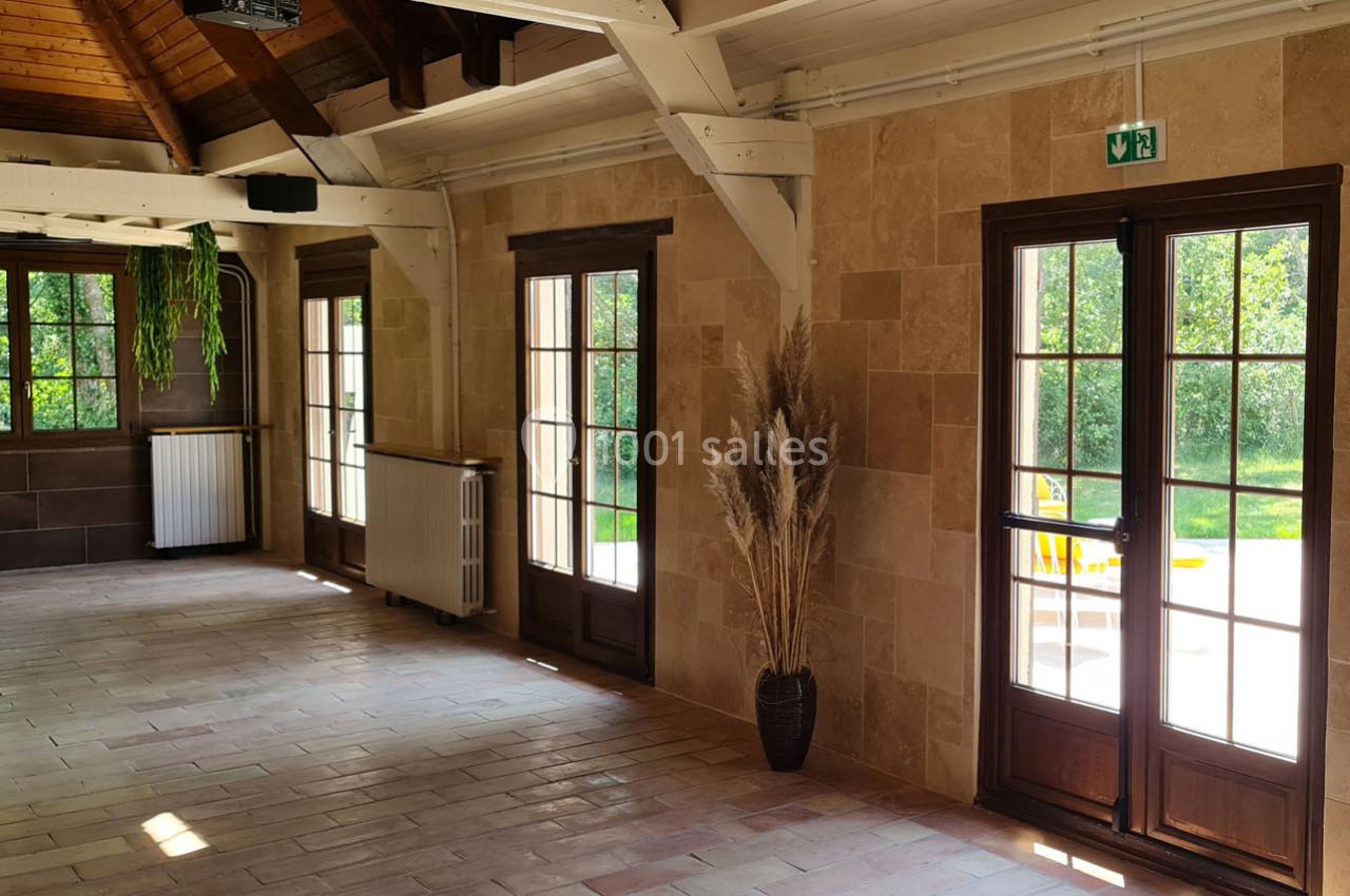 Salle lumineuse avec murs en pierre, poutres apparentes, grandes fenêtres vitrées et vue sur un jardin verdoyant.