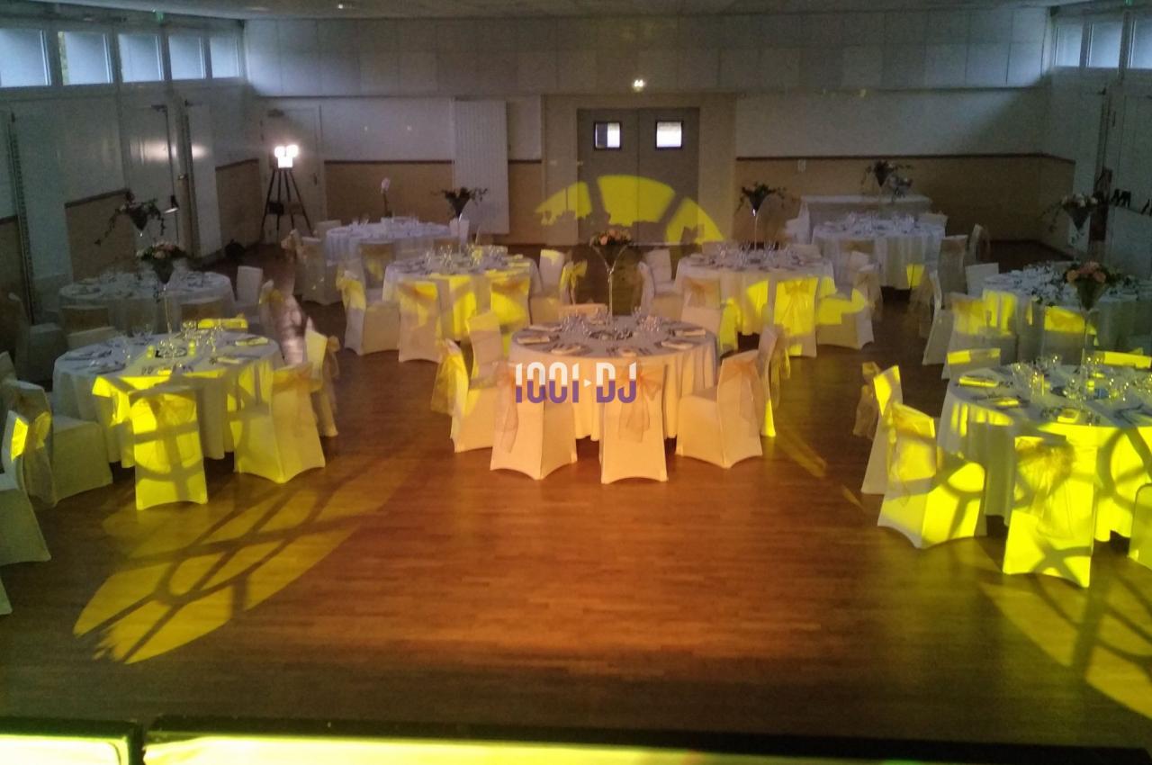 Salle de réception décorée avec des tables rondes dressées, éclairage jaune diffus et parquet en bois.