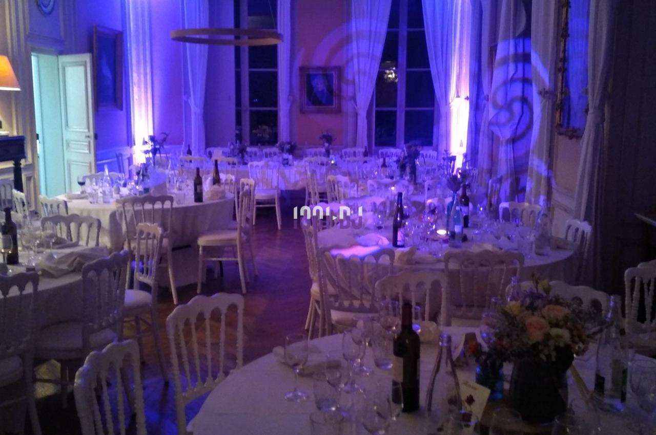 Salle de réception éclairée en bleu avec tables rondes dressées, chaises blanches et décorations élégantes.