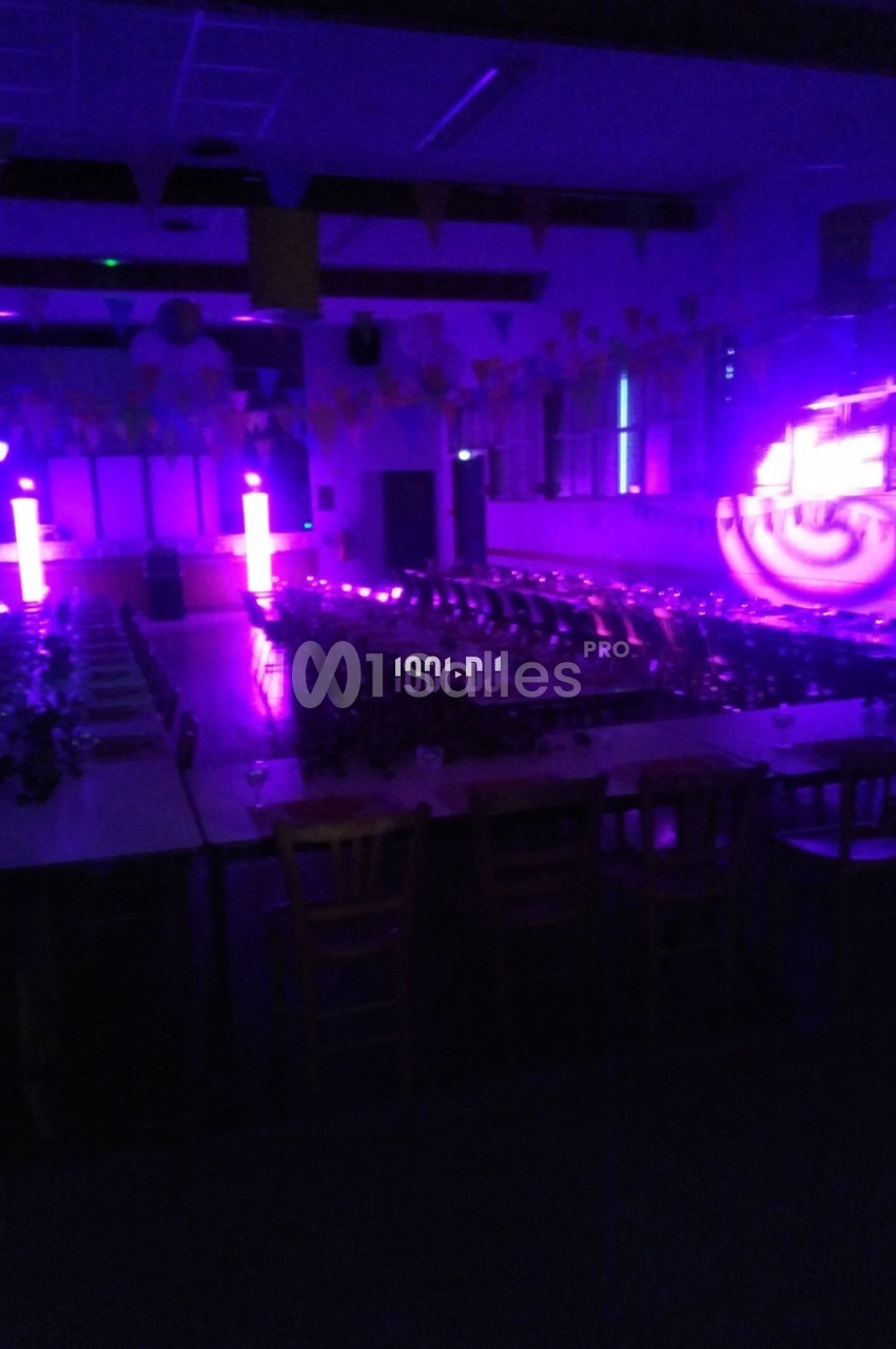 Salle sombre avec tables et chaises alignées, éclairée par des lumières violettes et des projecteurs lumineux.
