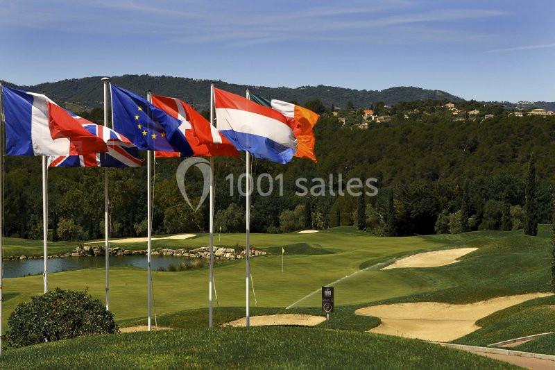 Drapeaux de plusieurs pays flottant devant un parcours de golf verdoyant avec des collines en arrière-plan.
