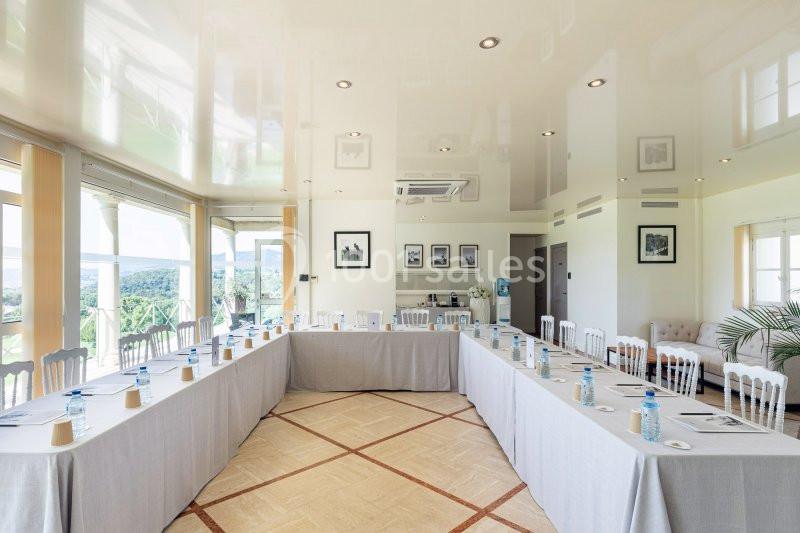 Salle de réunion lumineuse avec tables disposées en U, chaises blanches, bouteilles d'eau et vue sur l'extérieur.