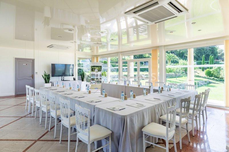 Salle de réunion lumineuse avec une grande table rectangulaire, des chaises blanches et vue sur un jardin verdoyant.