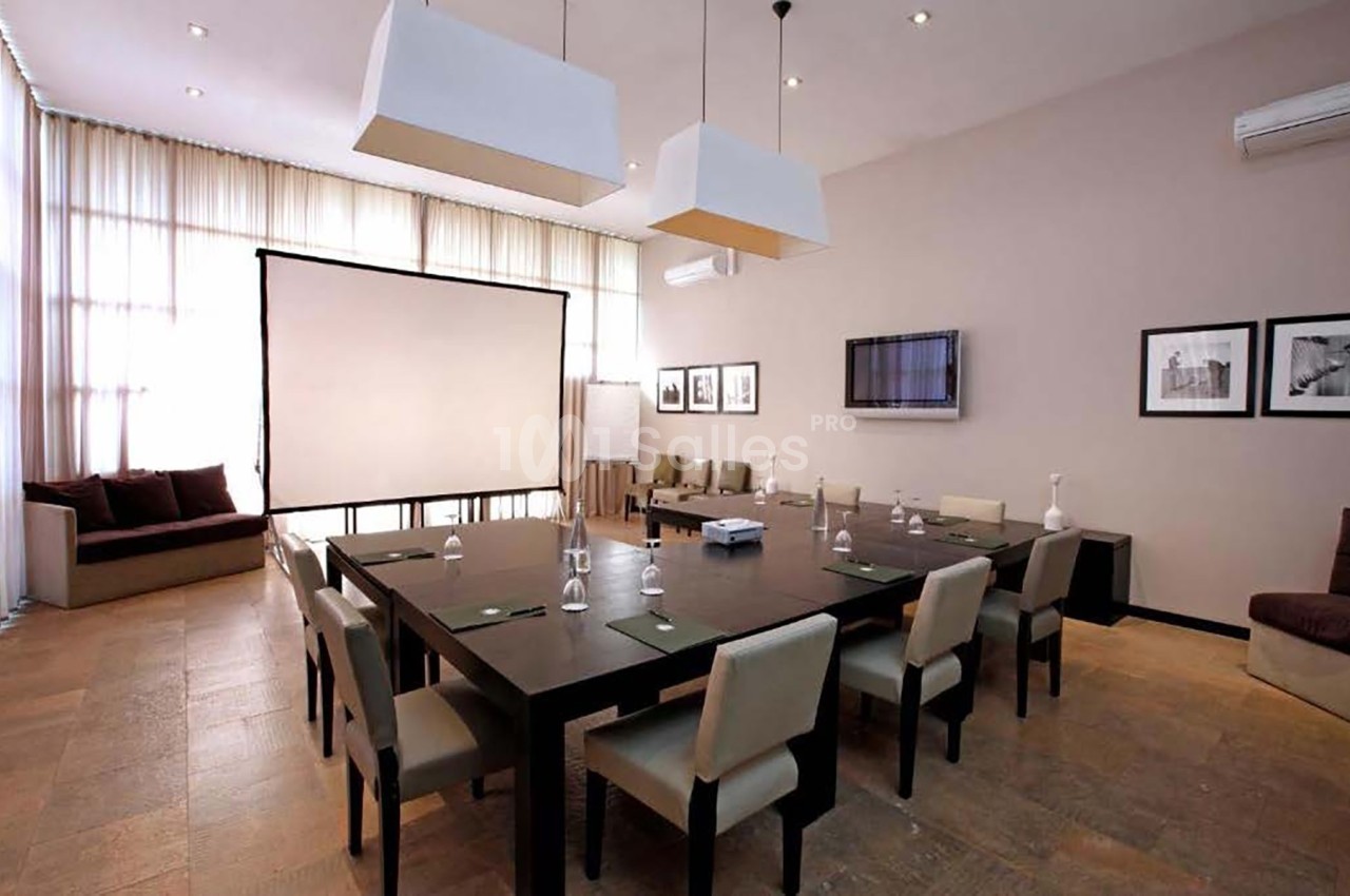 Salle de réunion lumineuse avec une grande table, des chaises, un écran de projection et des cadres accrochés au mur.