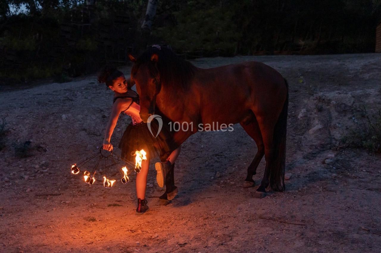 Une femme en jupe noire manipule un accessoire enflammé près d'un cheval brun dans un environnement extérieur sombre.