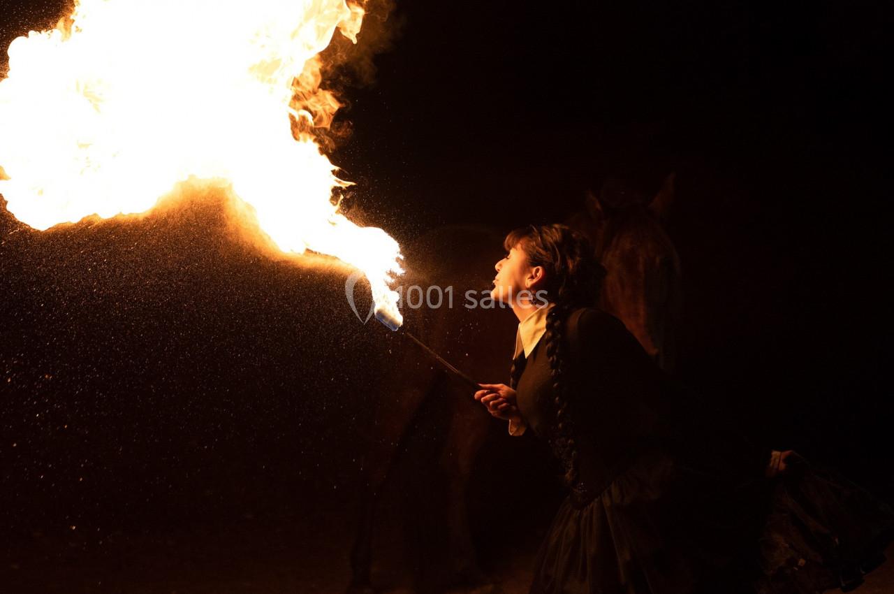 Une femme crache du feu dans l'obscurité, tenant une torche allumée, avec un cheval en arrière-plan.