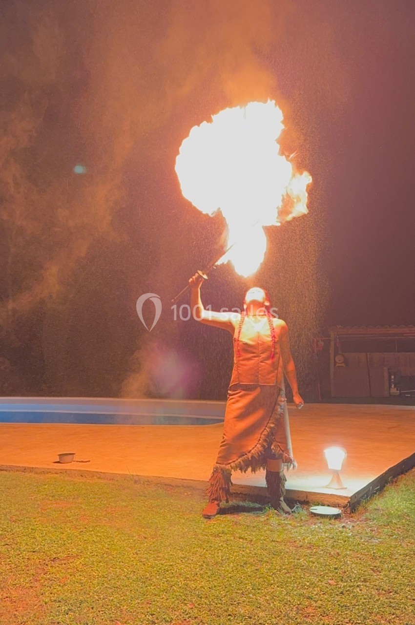 Une personne vêtue de tenue traditionnelle souffle du feu avec une torche dans un espace extérieur nocturne.