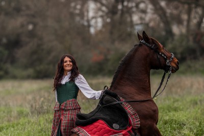 Femme en tenue traditionnelle caressant un cheval brun harnaché, dans un environnement naturel verdoyant.