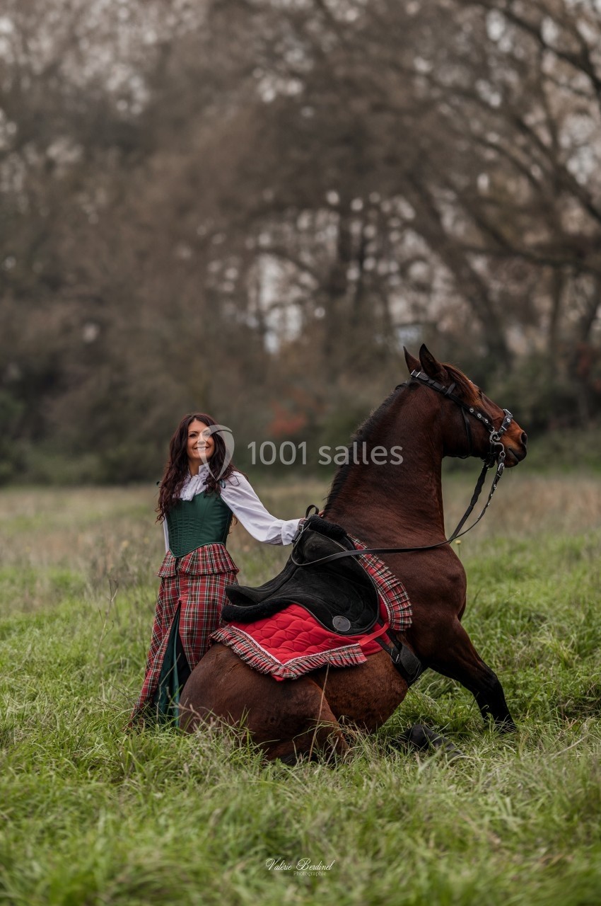 Une femme en tenue traditionnelle pose à côté d'un cheval assis dans un champ verdoyant.