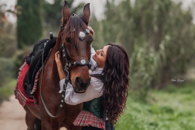 Femme en tenue traditionnelle caressant un cheval brun harnaché, dans un environnement naturel verdoyant.