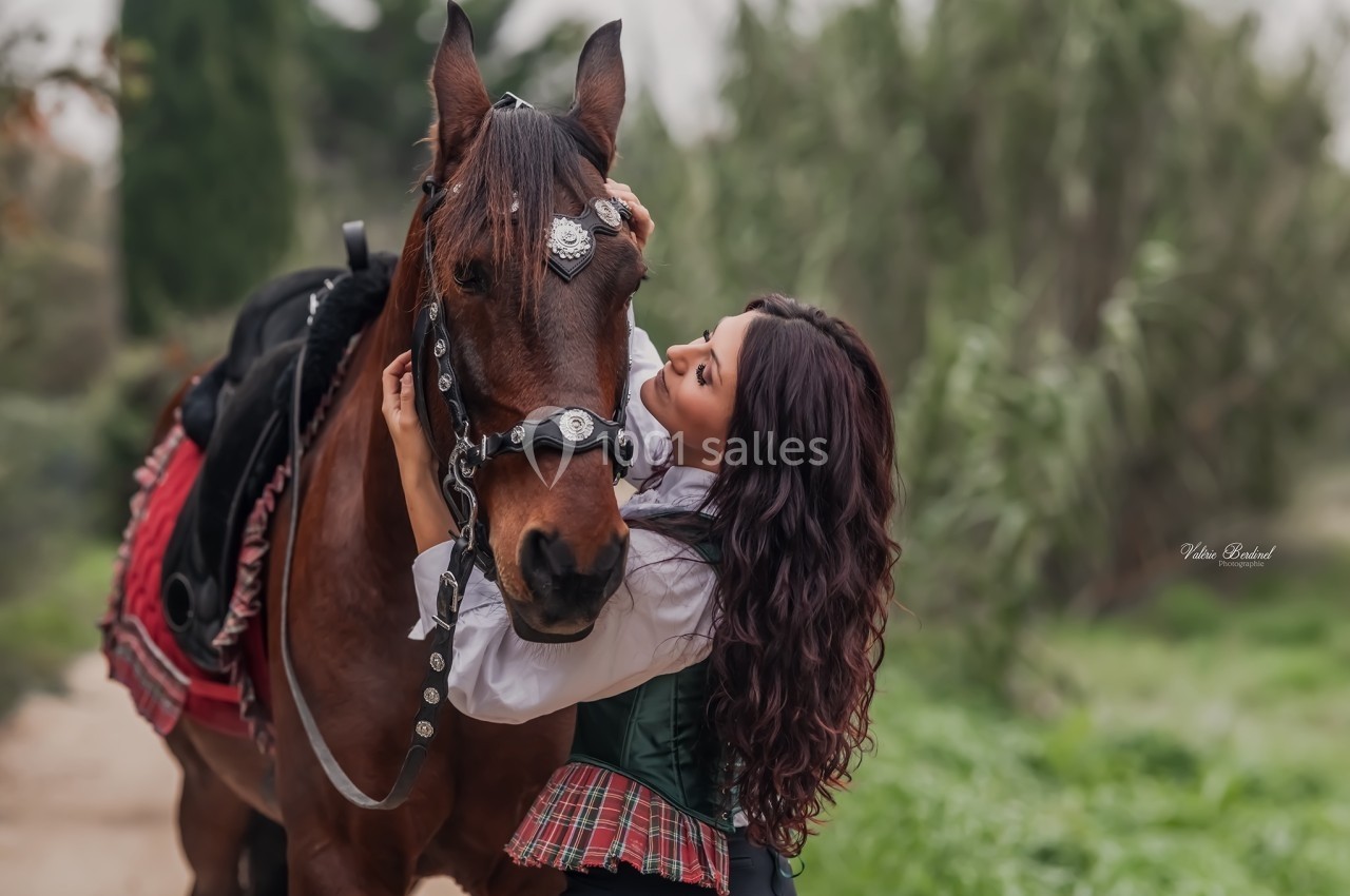 Femme en tenue traditionnelle caressant un cheval brun harnaché, dans un environnement naturel verdoyant.