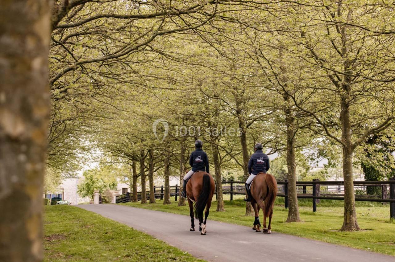 Deux cavaliers à cheval se promènent sur une allée bordée d'arbres dans un cadre verdoyant.