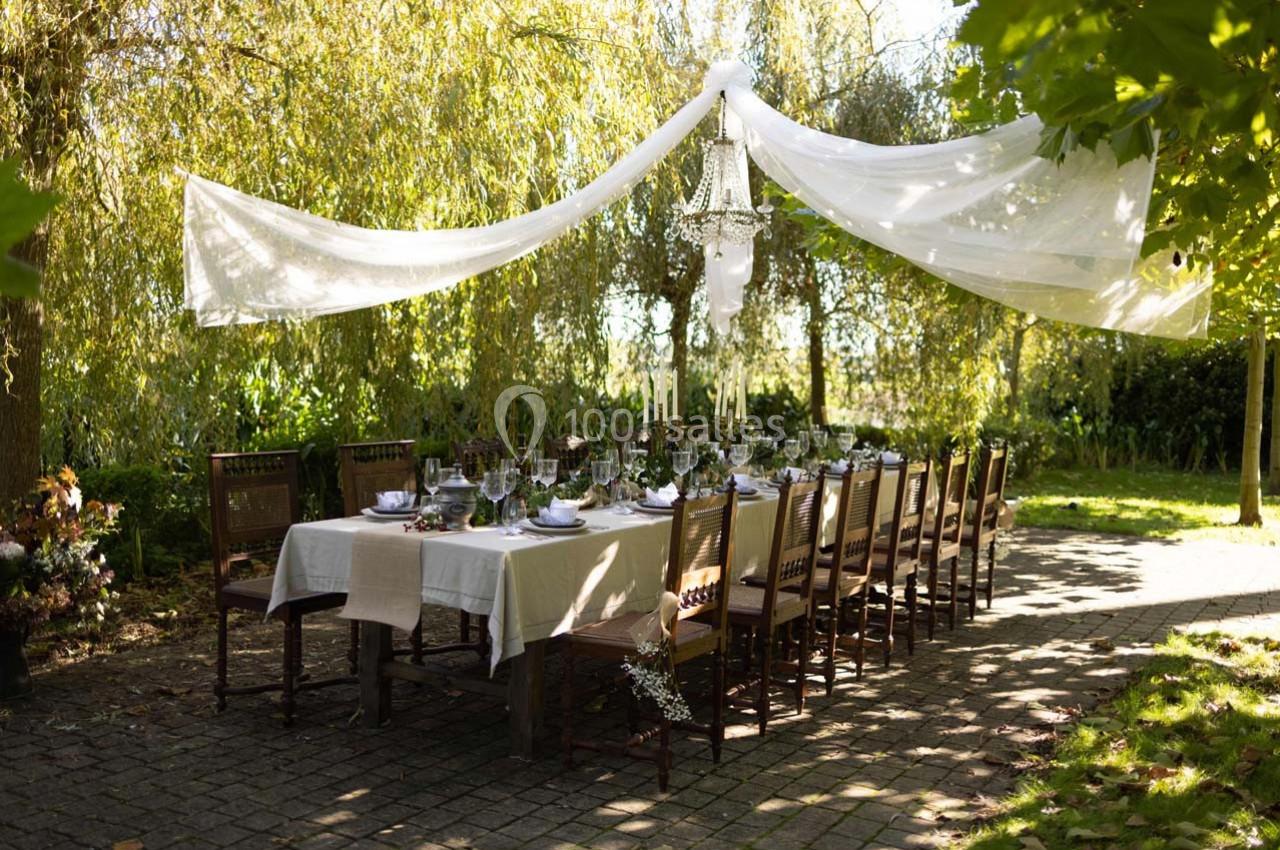 Table dressée pour un repas en extérieur sous un voile blanc, entourée de verdure et éclairée par un lustre suspendu.