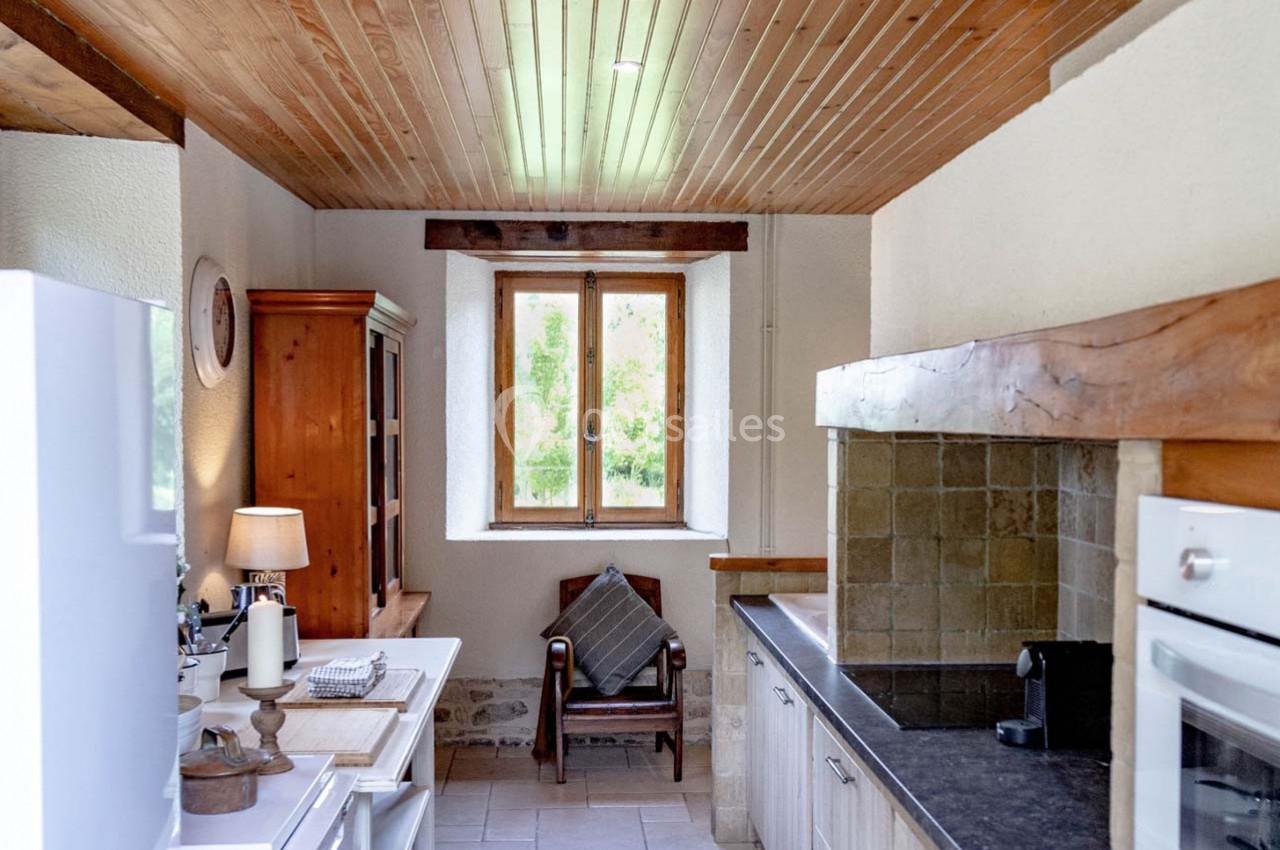 Cuisine lumineuse avec plafond en bois, fenêtre donnant sur un jardin, meubles rustiques et plan de travail en pierre.