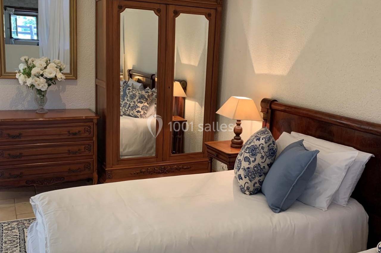 Deux lits simples avec linge blanc, coussins décoratifs bleus, armoire en bois avec miroir et lampe de chevet allumée.