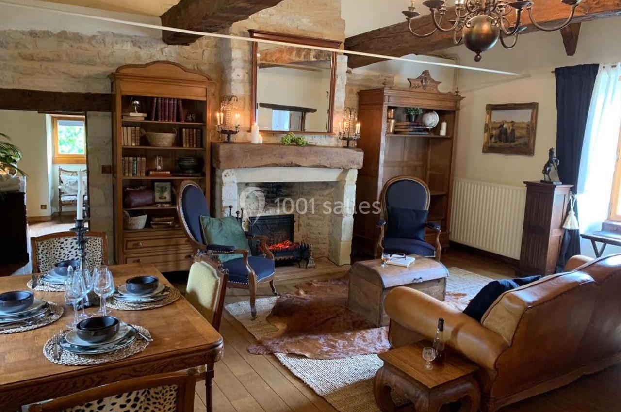 Salon rustique avec poutres apparentes, cheminée en pierre, meubles en bois et coin repas avec table dressée.