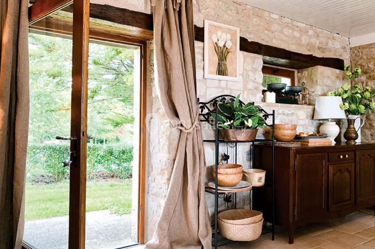 Intérieur rustique avec mur en pierre, meubles en bois, plantes et vue sur un jardin par une porte vitrée.
