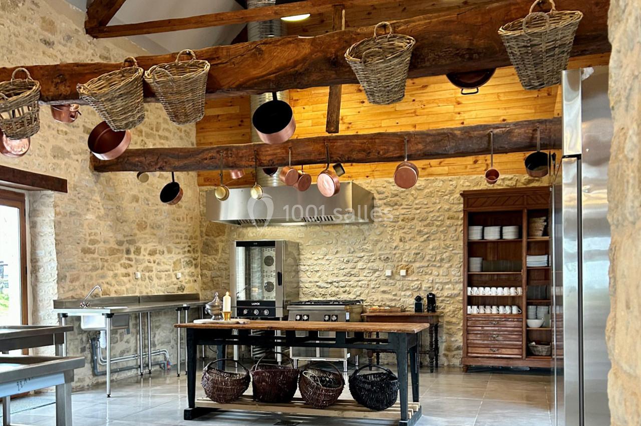 Cuisine rustique avec murs en pierre, poutres en bois, paniers suspendus et équipements modernes en inox.