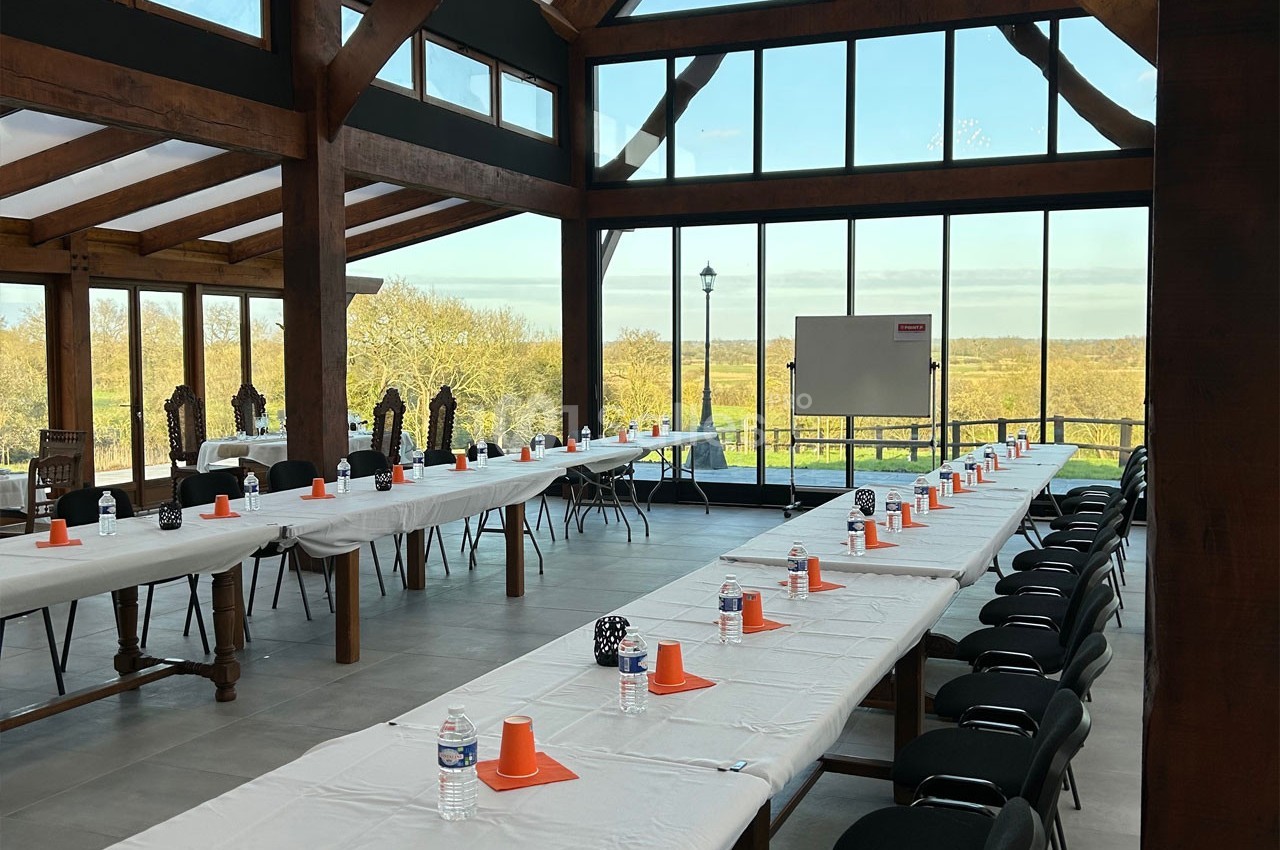 Grande salle lumineuse avec de longues tables dressées, chaises alignées, et vue sur un paysage extérieur verdoyant.