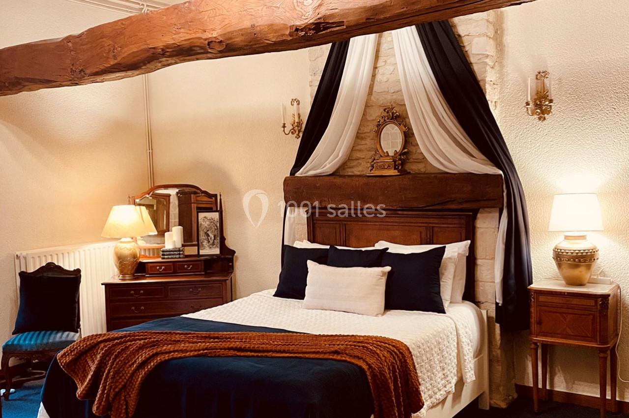 Chambre chaleureuse avec lit double, poutre apparente, draperies décoratives, meubles anciens et éclairage doux.