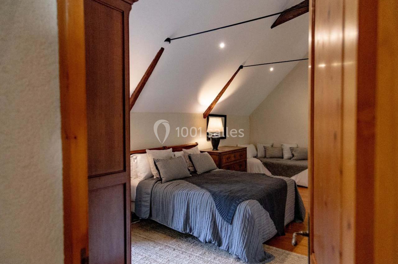 Chambre mansardée avec lit double, couvre-lit gris, lampe de chevet allumée et poutres apparentes au plafond.