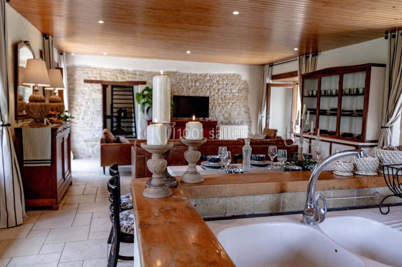 Cuisine ouverte avec évier en premier plan, vue sur un salon chaleureux avec murs en pierre et plafond en bois.