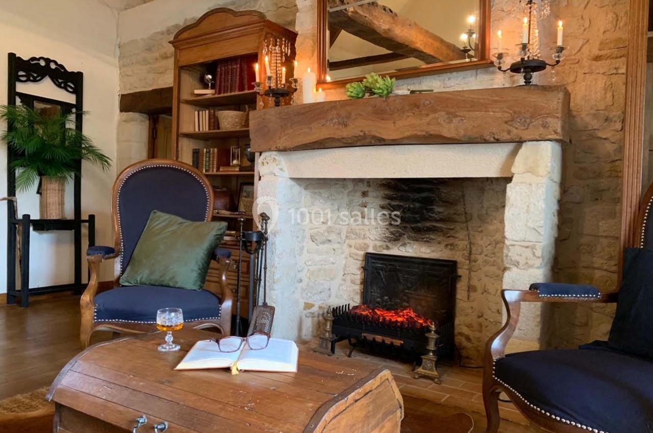 Salon chaleureux avec cheminée en pierre, fauteuils en bois, table basse en bois et étagères remplies de livres.