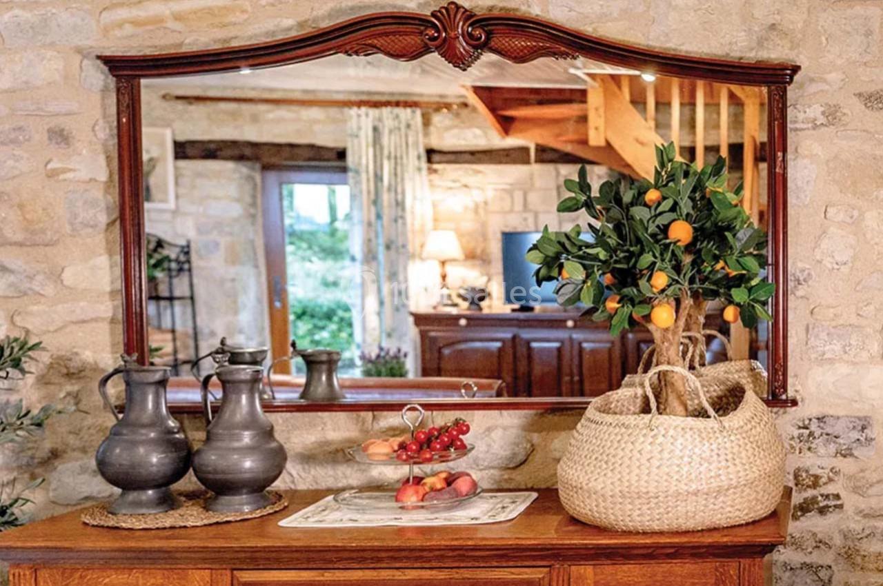 Un miroir encadré en bois reflète un intérieur rustique avec des murs en pierre, des objets décoratifs et une plante.