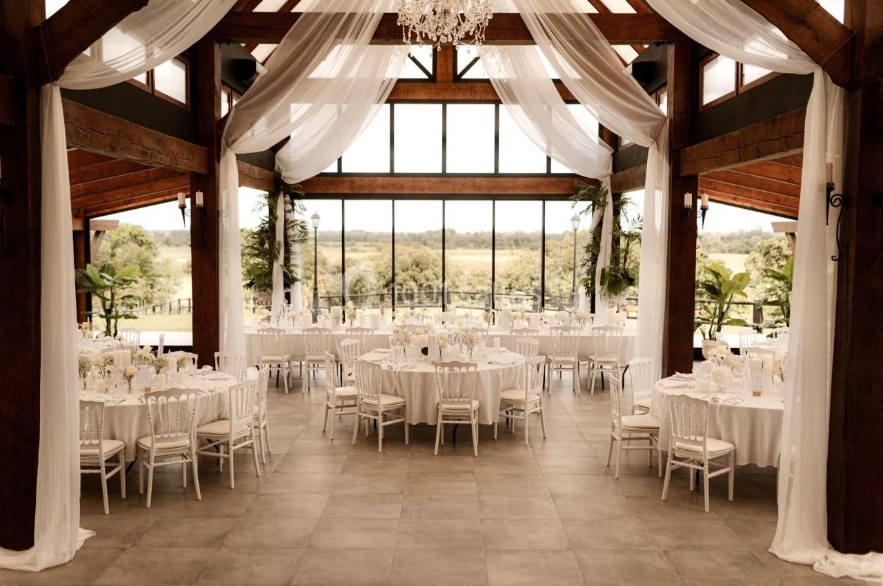 Salle de réception lumineuse avec tables rondes dressées, chaises blanches et grandes baies vitrées donnant sur un paysage…