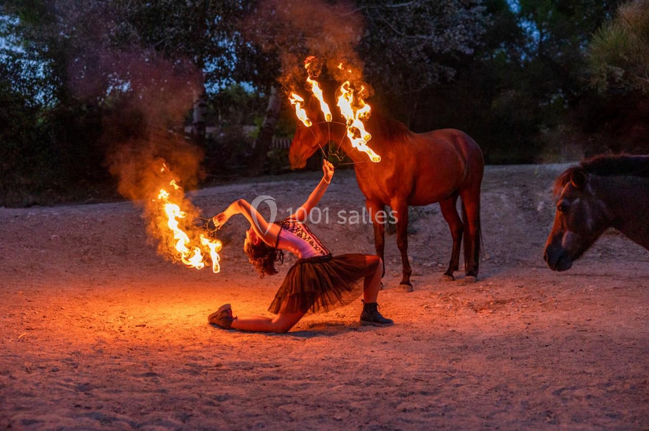 Une artiste manipule des torches enflammées près de chevaux dans un décor extérieur au crépuscule.