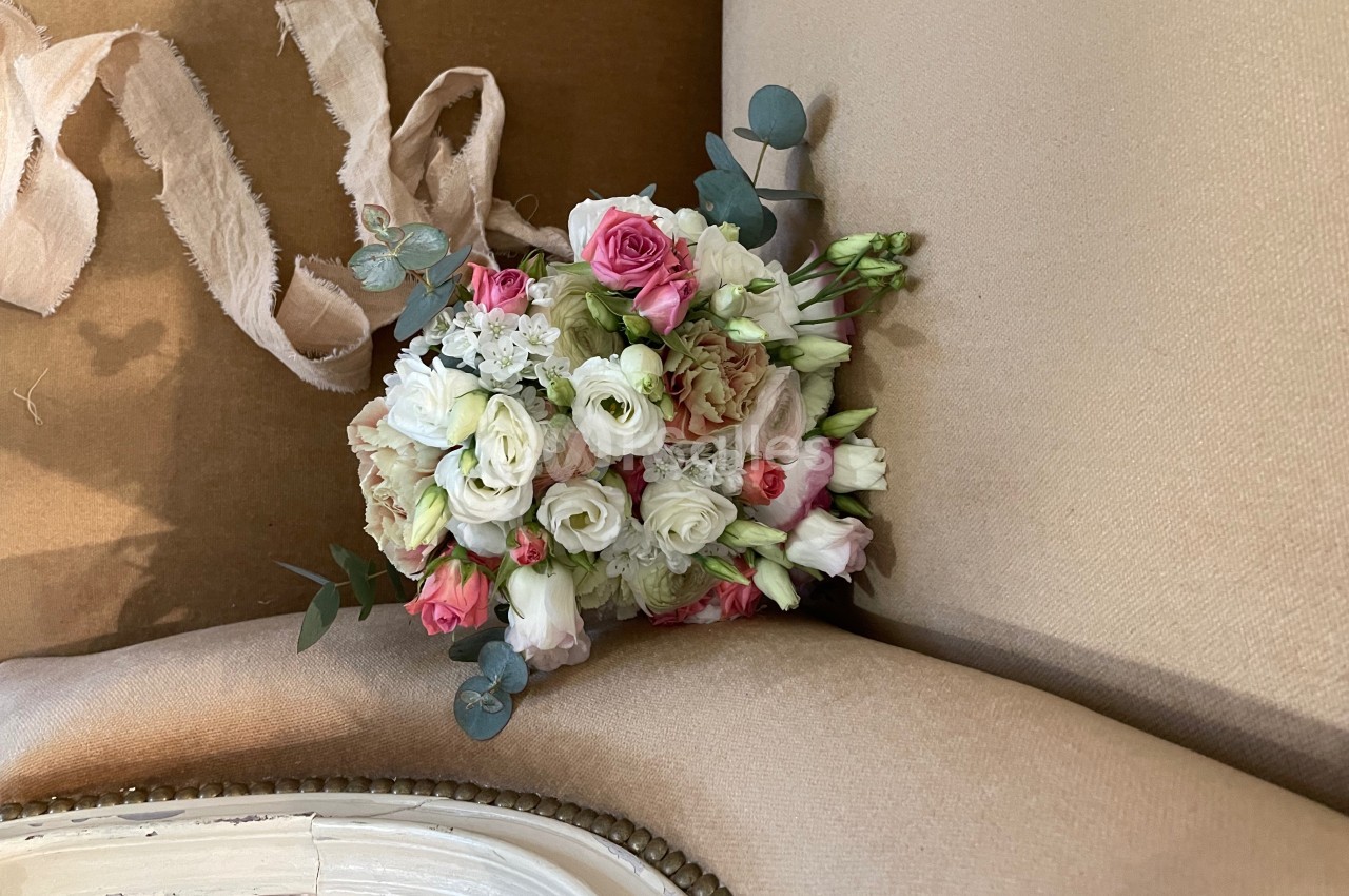 Bouquet de fleurs pastel composé de roses, renoncules et eucalyptus, posé sur un fauteuil beige.
