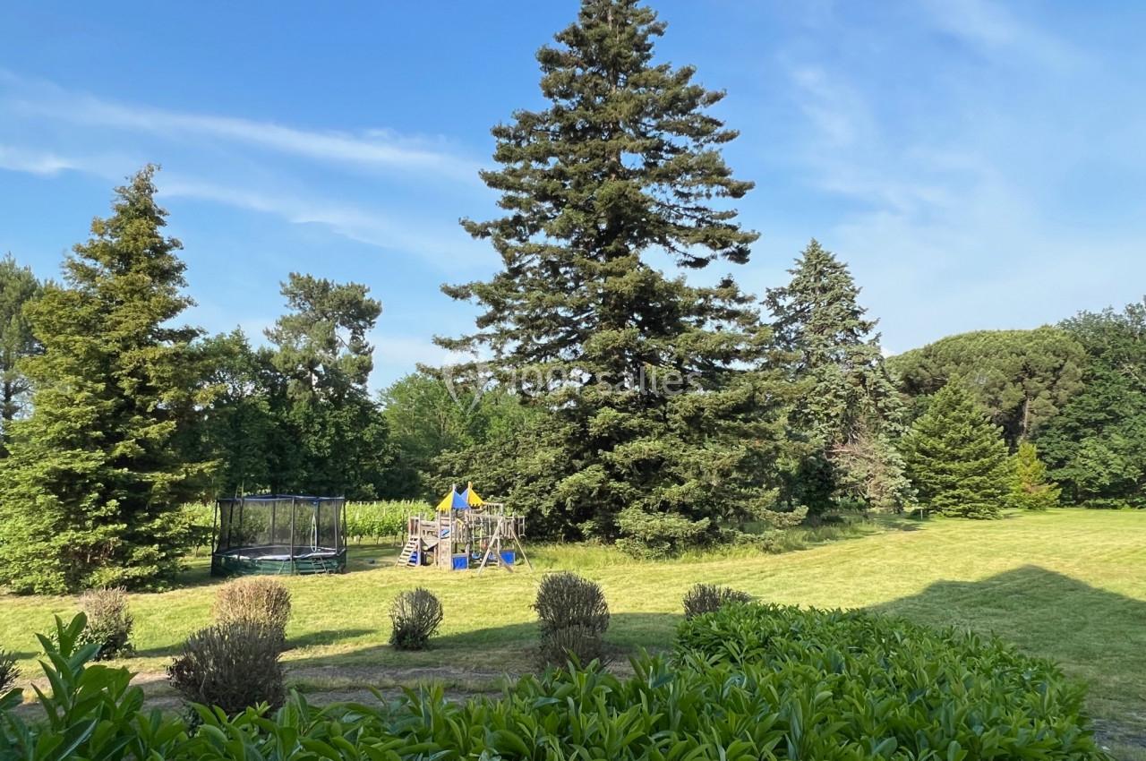 Grand jardin avec pelouse, arbres, trampoline et structure de jeu pour enfants sous un ciel dégagé.
