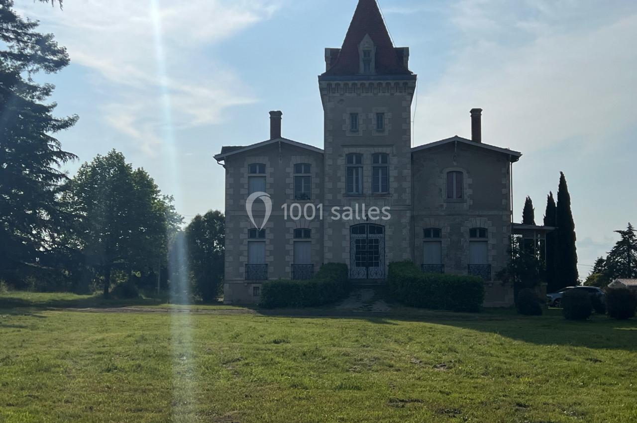 Grande maison en pierre avec une tour centrale et un toit rouge, entourée de verdure sous un ciel partiellement ensoleillé.