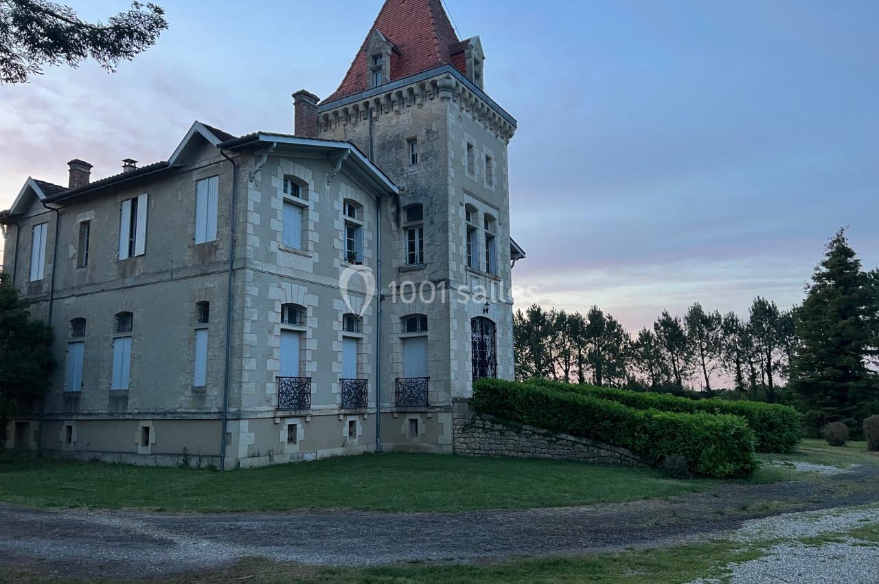 Grande maison en pierre avec une tour, entourée de verdure, sous un ciel légèrement nuageux au crépuscule.