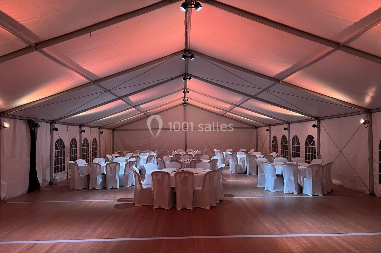 Intérieur d'une tente événementielle éclairée, avec des tables rondes dressées et des chaises blanches disposées autour.