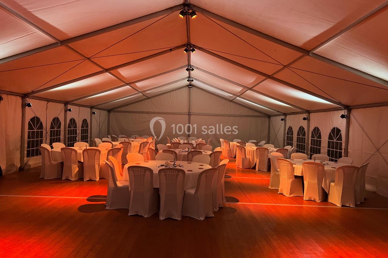 Salle sous chapiteau avec tables rondes dressées et chaises blanches, éclairée par une lumière chaude.