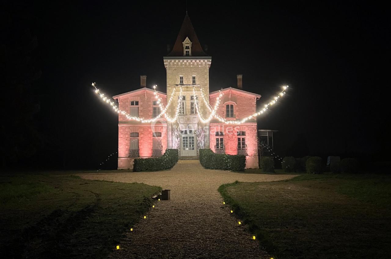 Façade d'un château éclairée par des guirlandes lumineuses, avec un chemin bordé de petites lumières sous un ciel nocturne.