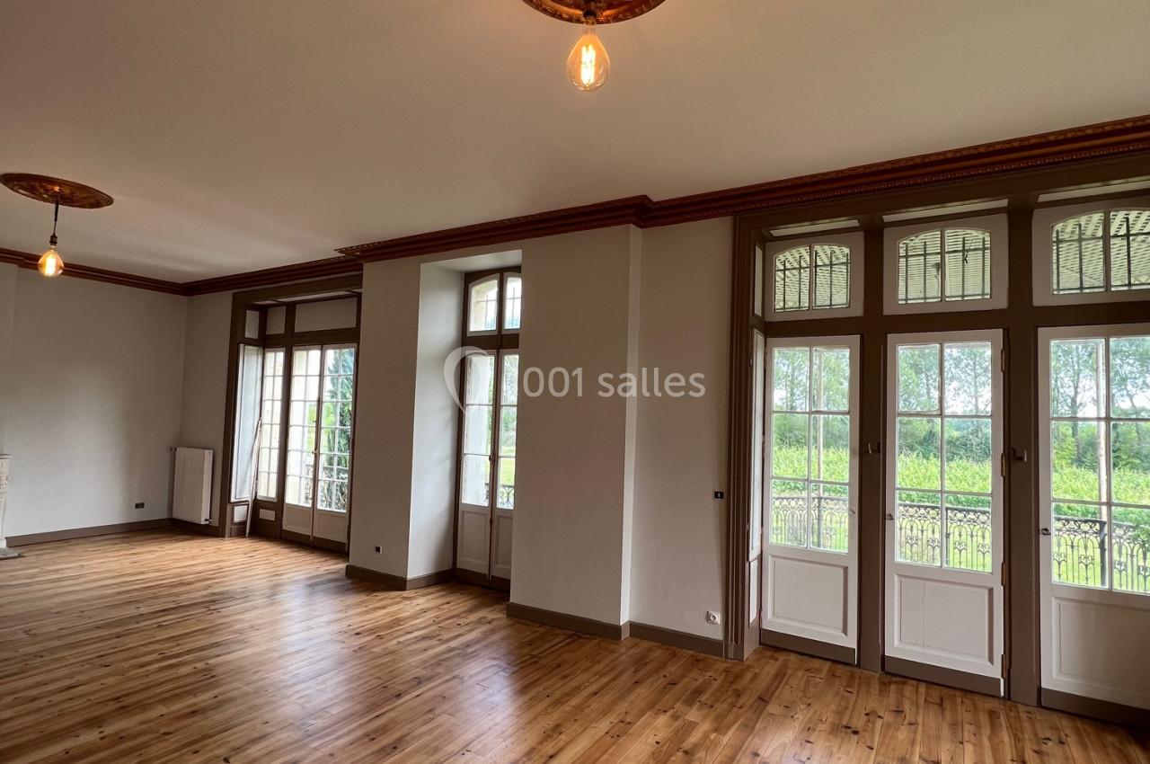 Salle lumineuse avec parquet en bois, moulures au plafond et grandes fenêtres donnant sur un espace vert.