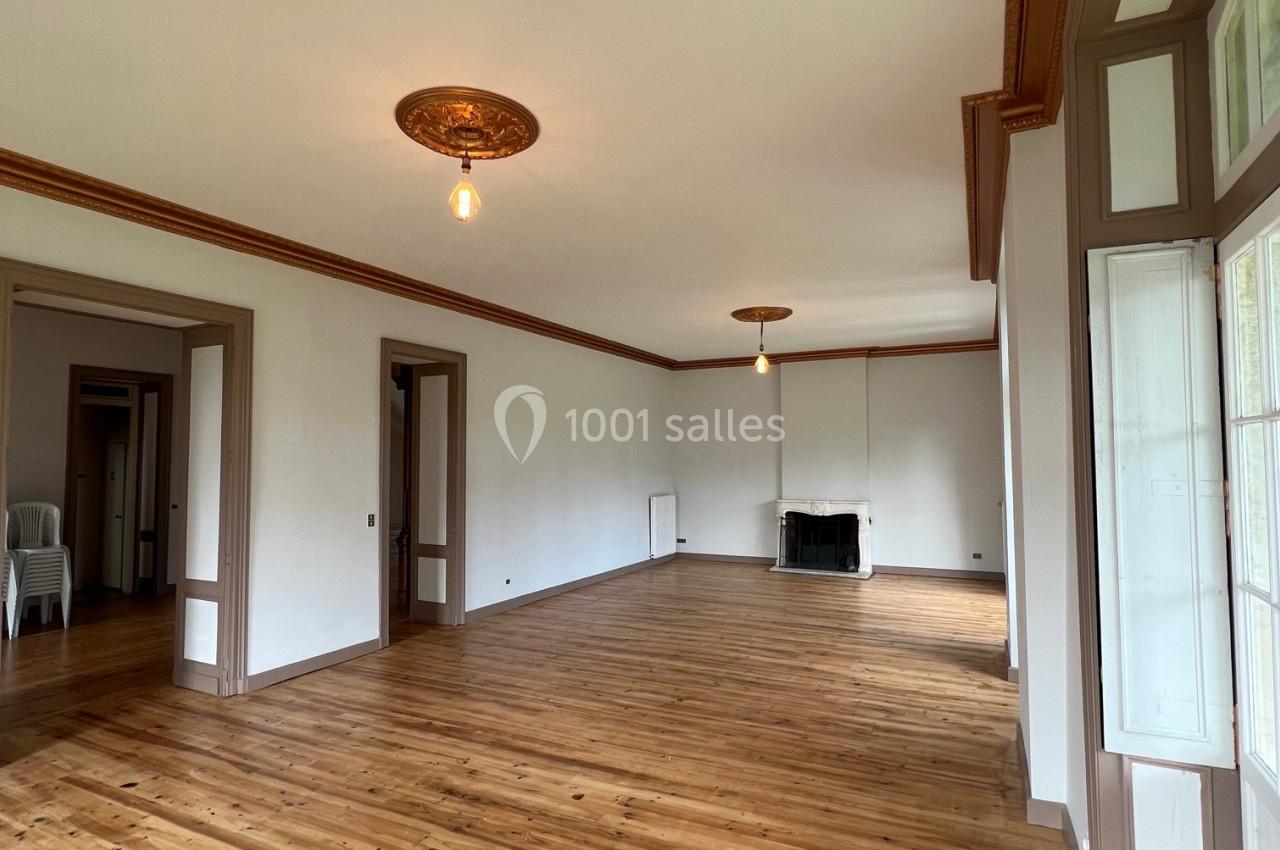 Grande pièce lumineuse avec parquet en bois, moulures au plafond, cheminée et grandes fenêtres donnant sur l'extérieur.