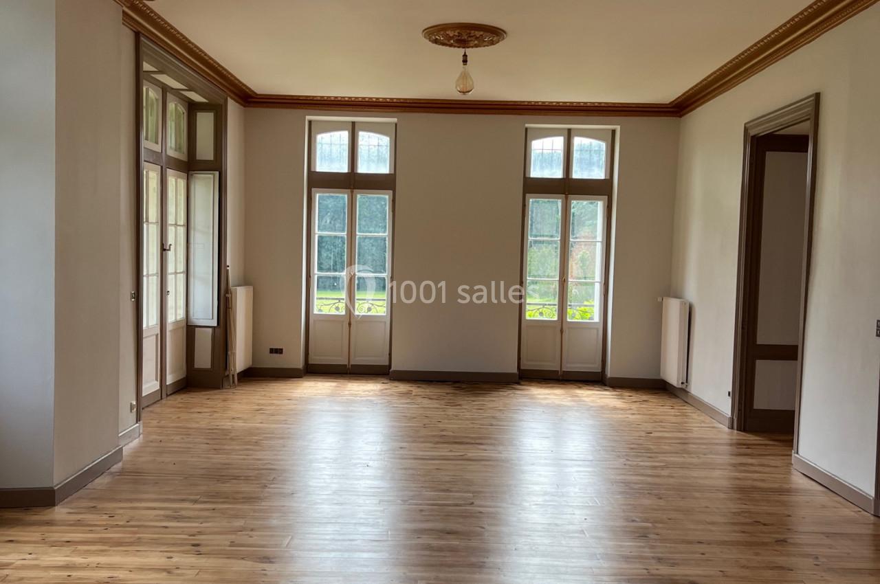 Salle lumineuse avec parquet en bois, grandes fenêtres à double battant et moulures au plafond.
