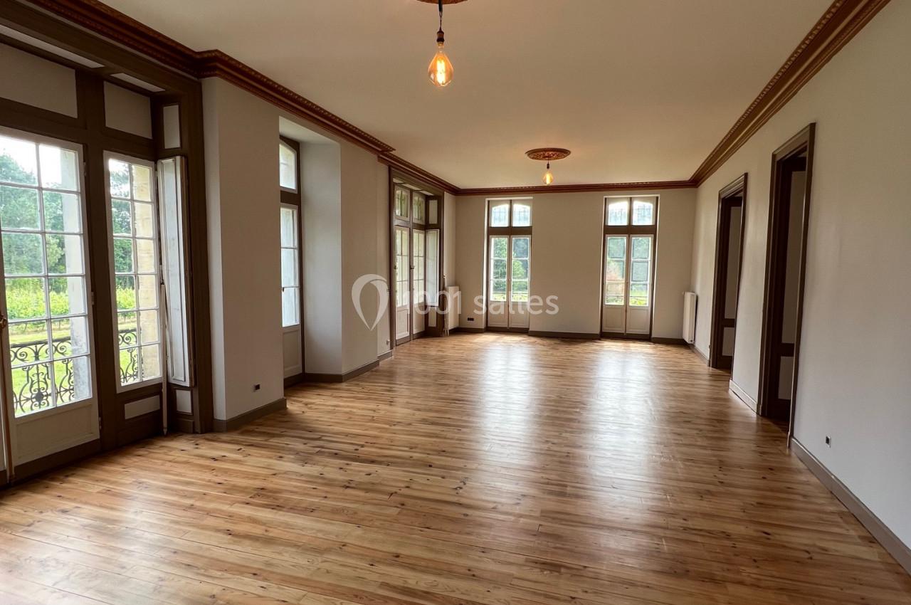 Salle lumineuse avec parquet en bois clair, grandes fenêtres et moulures au plafond, donnant sur un espace verdoyant.