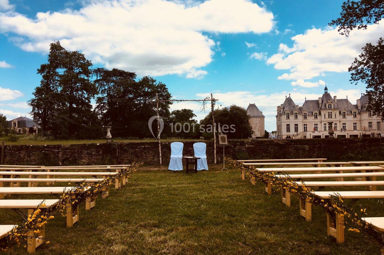 Allée de bancs en bois décorés menant à une arche nuptiale, avec un château en arrière-plan sous un ciel partiellement…