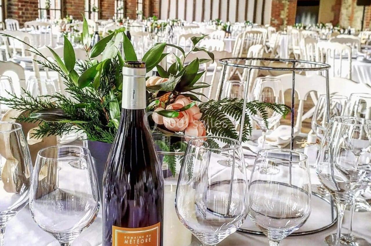 Salle de réception élégante avec tables dressées, centre de table floral et bouteille de vin au premier plan.